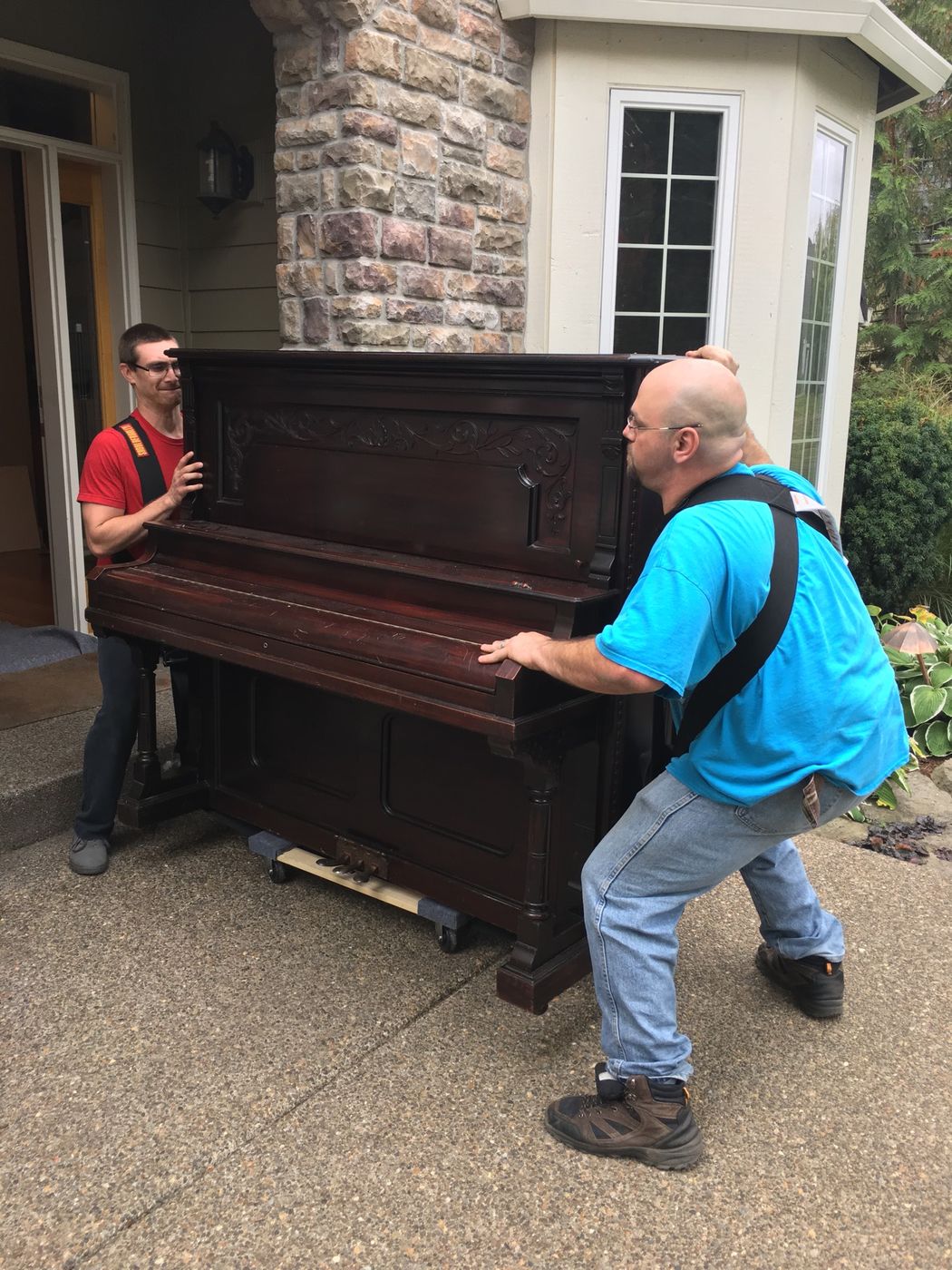 Two movers walking an upright piano out of a front door
